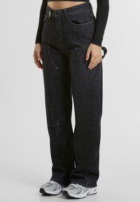 Pantaloni a gamba larga in denim nero con vita alta, cuciture in evidenza, tasche laterali e chiusura a bottone, abbinati a sneakers grigie.