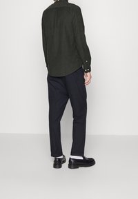 Ciemnozielona koszula na guziki z długimi rękawami, z tylną wstawką, w połączeniu z luźnymi czarnymi spodniami i czarnymi butami na masywnych podeszwach.