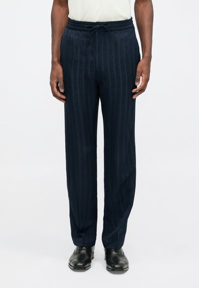 Tamsiai mėlynos dryžuotos kelnės su elastingu juosmeniu ir raišteliu, pagamintos iš lengvos medžiagos, derinamos su juodais bateliais.