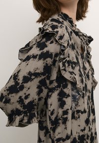 Blusa de patrón negro y beis con acentos fruncidos, corte holgado, escote tipo ojal y mangas con puños ajustados. Tejido suave con una apariencia texturizada.
