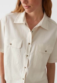 Camicia a maniche corte in cotone color beige chiaro con pettorina abbottonata, due tasche sul petto e dettagli in bottone argentato.