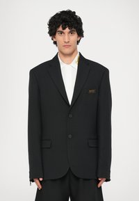 Črn oversize blazer iz teksturirane tkanine, opremljen z dvema gumboma, stranskimi žepi in majhnim blagovnoznamčnim znakom na prsih.