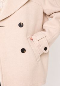 Cappotto in misto lana beige con design doppiopetto, bottoni neri e tasca laterale. Presenta un colletto strutturato e pattini delle maniche regolabili.