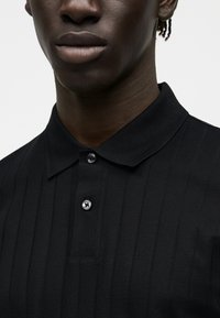 Κοντινό πλάνο ενός ατόμου που φορά ένα μαύρο ριμπ πικέ με γιακά και δύο ορατά κουμπιά, εμφανίζοντας το κάτω μέρος του προσώπου και τον λαιμό.