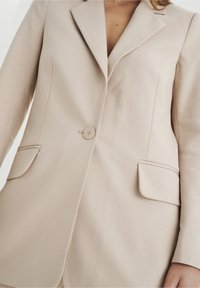 Blazer beige ajustado con solapa muesca, cierre de un solo botón y dos bolsillos frontales. Textura suave, con forro en contraste.