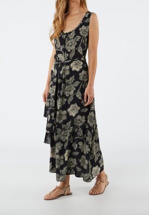Kobieta ubrana w czarną, bezrękawnikową sukienkę maxi w beżowy kwiatowy wzór z pasującym paskiem, do tego beżowe płaskie sandały z koralikami.