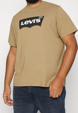 Skægget person iført en tanfarvet Levi's t-shirt og mørkeblå jeans, stående med den ene hånd ved siden af sig og den anden nær sin lomme.