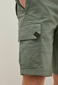 Shorts cargo verts avec plusieurs poches, coutures surpiquées et une étiquette noire portant un logo de couronne, mettant en valeur un tissu durable et texturé.
