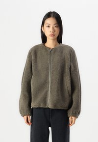 Oliivivihreä fleece-takki, jossa on pyöreä kaulus, etuvetoketju ja sivutasku. Takissa on teksturoitu, pörröinen pinta ja elastiset hihansuut.