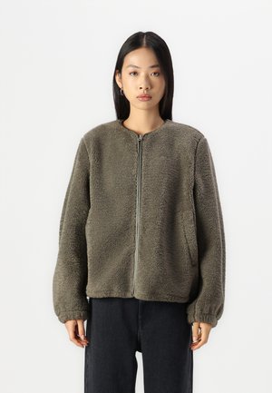Oliivivihreä fleece-takki, jossa on pyöreä kaulus, etuvetoketju ja sivutasku. Takissa on teksturoitu, pörröinen pinta ja elastiset hihansuut.