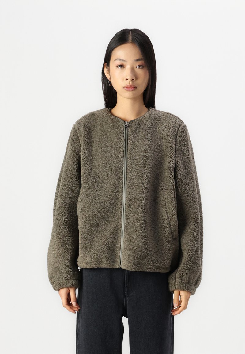 Oliivivihreä fleece-takki, jossa on pyöreä kaulus, etuvetoketju ja sivutasku. Takissa on teksturoitu, pörröinen pinta ja elastiset hihansuut.