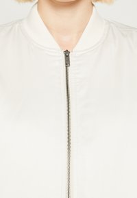 Lehká bílá bomber bunda s žebrovaným límcem a stříbrným zipem. Materiál má hladkou texturu a minimalistický design.