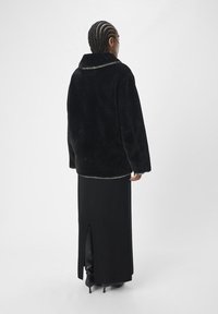 Zwarte neplederen jas met een grote kraag en zilveren hardware details langs de hals. Draagt over een zwarte, lange rok met een rugsplit.
