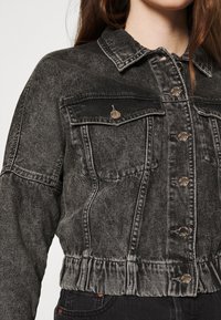 Bledě černá džínová bunda se stříbrnými knoflíky, klopovými náprsními kapsami, elastickým pasem, nošena přes černé kalhoty.