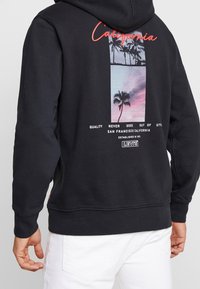 Sudadera con capucha negra que presenta un estampado gráfico de palmeras y un atardecer en la parte posterior, con texto rojo y acentos de marca en blanco.