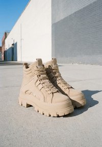 Paire de bottes beige à lacets avec semelles épaisses posées sur un sol en béton près de murs en briques claires à l'extérieur.