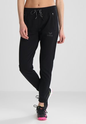 Personne portant un pantalon de sport noir avec des poches zippées et une taille à cordon, associé à des chaussures de sport noires et roses.