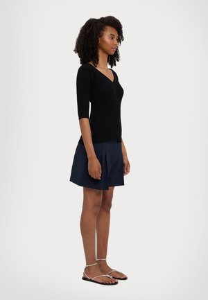 Zwarte V-hals gebreide top met driekwartmouwen, gecombineerd met een donkerblauwe plissérok en zilveren platte sandalen. Eenvoudige, casual silhouet.