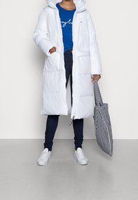 Persona que lleva un abrigo de plumas largo y blanco, una camisa azul, jeans oscuros, zapatillas blancas, y sostiene una bolsa de tote a rayas blancas y negras.