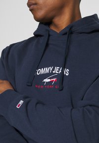 Marinblå hoodie i bomull med en broderad "TOMMY JEANS"-logotyp och röda detaljer. Har en dragsko i huven och ribbade ärmslut.