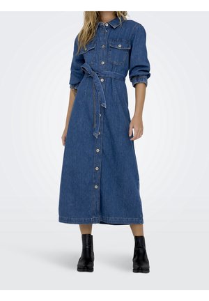 Kvinde iført en langærmet blå denim-knap-front kjole med brystlommer, et taljebælte og sorte ankelstøvler, stående mod en ensfarvet baggrund.