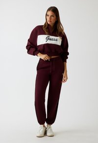 Bordeaux sportieve trainingsuit met een witte strook waarop "Guess" in zwarte letters is geschreven; omvat een cropped sweatshirt en tapered broek met zwarte accenten.