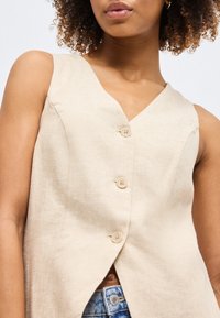 Femme portant un haut beige sans manches boutonné avec un col en V, associé à un jean en denim bleu, sur un fond blanc uni.