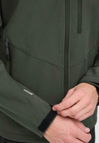 Des mains ajustant la sangle de la manche d'une veste softshell WP Pro 8000 vert foncé avec des détails noirs sur la fermeture éclair et les poches.