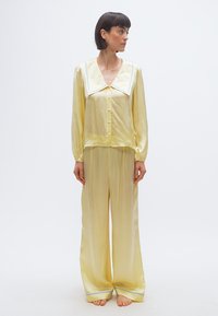 Γυναίκα που στέκεται ξυπόλητη φορώντας λαμπερό σετ πιτζάμες από απαλό κίτρινο σατέν με φαρδιά γιακά και μπλε περιθώριο, κοιτάζοντας προς τα δεξιά της.