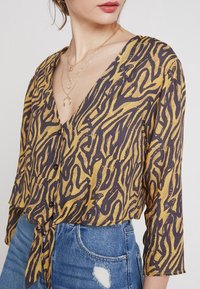 Yellow and dark brown patterned blouse with a V-neck, 3/4 sleeves, button closure, and a tie-front detail. Worn with high-waisted denim shorts.