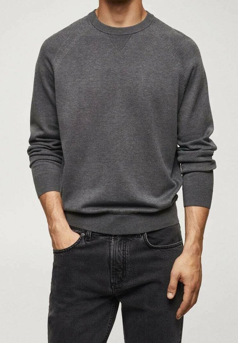 Homme portant un pull gris foncé à col rond avec manches raglan et un jean noir, debout avec une main dans la poche sur un fond clair.
