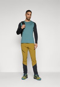 Herren Langarmshirt in Teal mit schwarzen Ärmeln, kombiniert mit gelb-braunen Hosen mit schwarzen Akzenten. Enthält einen Rucksack und Outdoor-Schuhe.