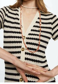 Schwarz-weiß gestreifter Strickcardigan mit großen Knöpfen, kombiniert mit einer bunten Perlenkette mit Muscheln und einem gemusterten Textur.