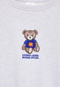 Hnedý plyšový medvedík v modrej košeli s oranžovou hviezdou, nad ktorým je na svetlosivej látke nápis "Roztomilosť nabitá. Trpezlivosť offline."