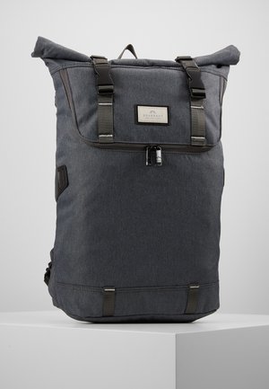 Sac à dos gris foncé à enroulement avec boucles noires, poche zippée à l'avant et patch logo rectangulaire sur une surface blanche unie.