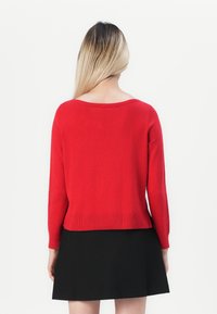 Žena s dlhými blond vlasmi, oblečená v červenom svetri s dlhým rukávom a čiernej sukni, otočená chrbtom proti bielemu pozadiu.