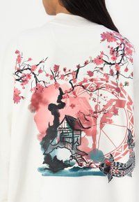 Fehér pulóver, amelyen egy nagy, színes virág- és tájnyomtatás található. Rózsaszín cseresznyevirágok, egy narancs motívum és egy részletes hal is szerepel rajta.