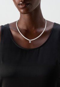 Collana di perle con perle bianche alternate e un accento di cristallo a forma di cuore, indossata sopra un top nero. Design classico e lucido.