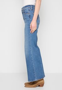 Femme portant un jean bleu taille haute, droit, et des bottines à talons couleur beige, debout devant un fond uni clair.