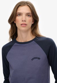 Młoda kobieta o jasnobrązowych falowanych włosach, ubrana w niebieski i granatowy top z raglanowymi rękawami z logo "SUPERDRY" na klatce piersiowej.