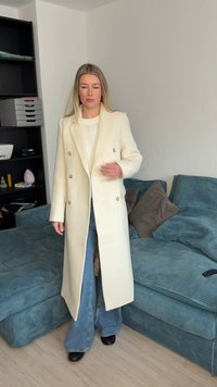 Γυναίκα που φοράει ένα μακρύ μπεζ παλτό, λευκό πουλόβερ και μπλε τζιν, στέκεται κοντά σε έναν τιρκουάζ καναπέ από κορδόνι με μαξιλάρια σε ένα μινιμαλιστικό σαλόνι.