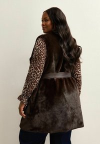 Brązowa kamizelka z sztucznego futra z paskiem w talii, noszona na długim rękawie koszuli w brązowo-beżowy leopardzi wzór, na neutralnym tle.