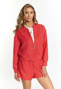 Rote, strukturierte Bomberjacke mit Strassakzenten und Reißverschlussfront. Kombiniert mit passenden Shorts, die einen elastischen Bund und Taschen haben.