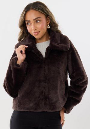 Mujer con chaqueta de piel sintética marrón oscuro con cuello ancho, pantalones negros, top claro y pendientes de aro dorados, mirando hacia un lado.