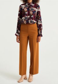 Blus med blommönster i mörkröd, blå och brun, matchad med högmidjade, vida byxor i senapsgul och beige blockklackskor.