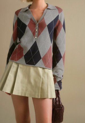 Femme portant un pull à manches longues à motif losanges avec des boutons et une mini-jupe plissée beige, tenant un sac à main tressé marron foncé.