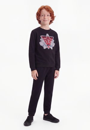 Garçon aux cheveux roux debout et souriant, portant un sweat-shirt noir avec un motif rouge et blanc, ainsi qu'un pantalon et des chaussures noirs assortis.