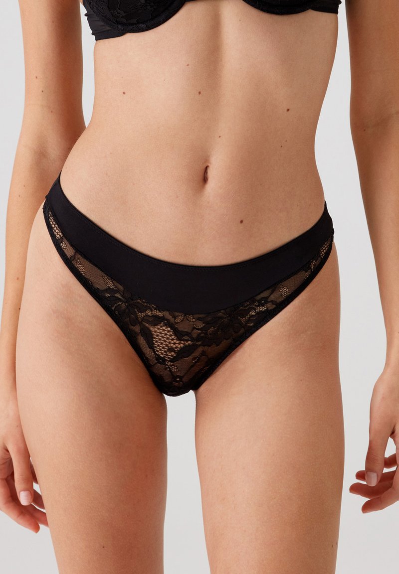 Zwarte kant thong met een gladde tailleband en een bloemenkantpatroon. Semi-doorzichtige stof met een contrasterende ondoorzichtige band bovenaan.
