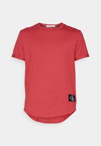 Camiseta de algodón roja con mangas cortas, cuello redondo, dobladillo curvado y un parche de logo negro en la parte inferior izquierda. Textura suave, estilo casual.