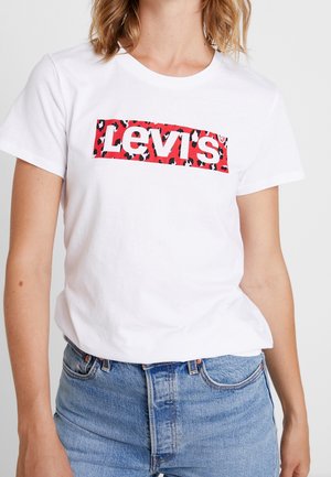 Kobieta nosząca białą koszulkę Levi's z czerwono-czarnym logo w panterkę, w połączeniu z jasnoniebieskimi dżinsami.